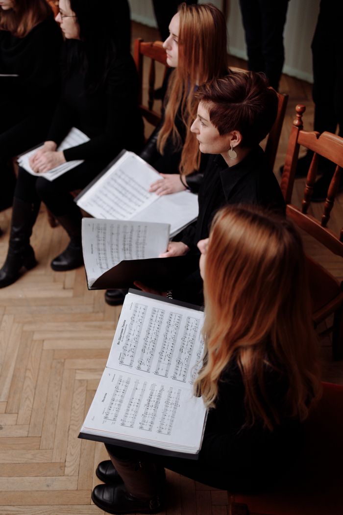The Art of Drawing Readers In: Your attractive post title goes here Group of women in a choir holding music sheets, seated on wooden chairs, practicing indoors.